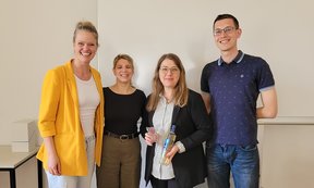 Zu sehen sind die Veranstalter_innen des Workshops. Sie stehen nebeneinander und lächeln. Eine der Personen hält ein kleines Geschenk in der Hand.