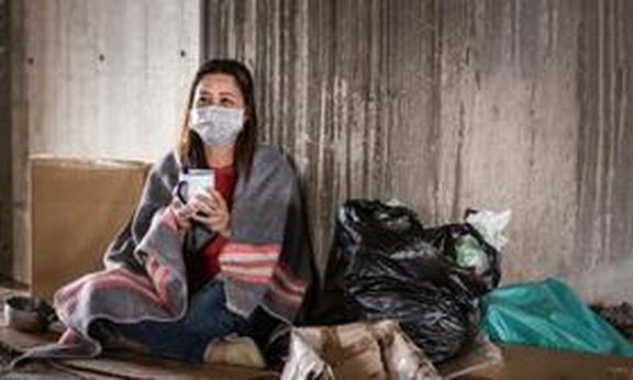 Person mit Mund-Nasen-Schutz sitz auf dem Boden in eine Deckel gewickelt mit gefüllten Müllsäcken neben sich.