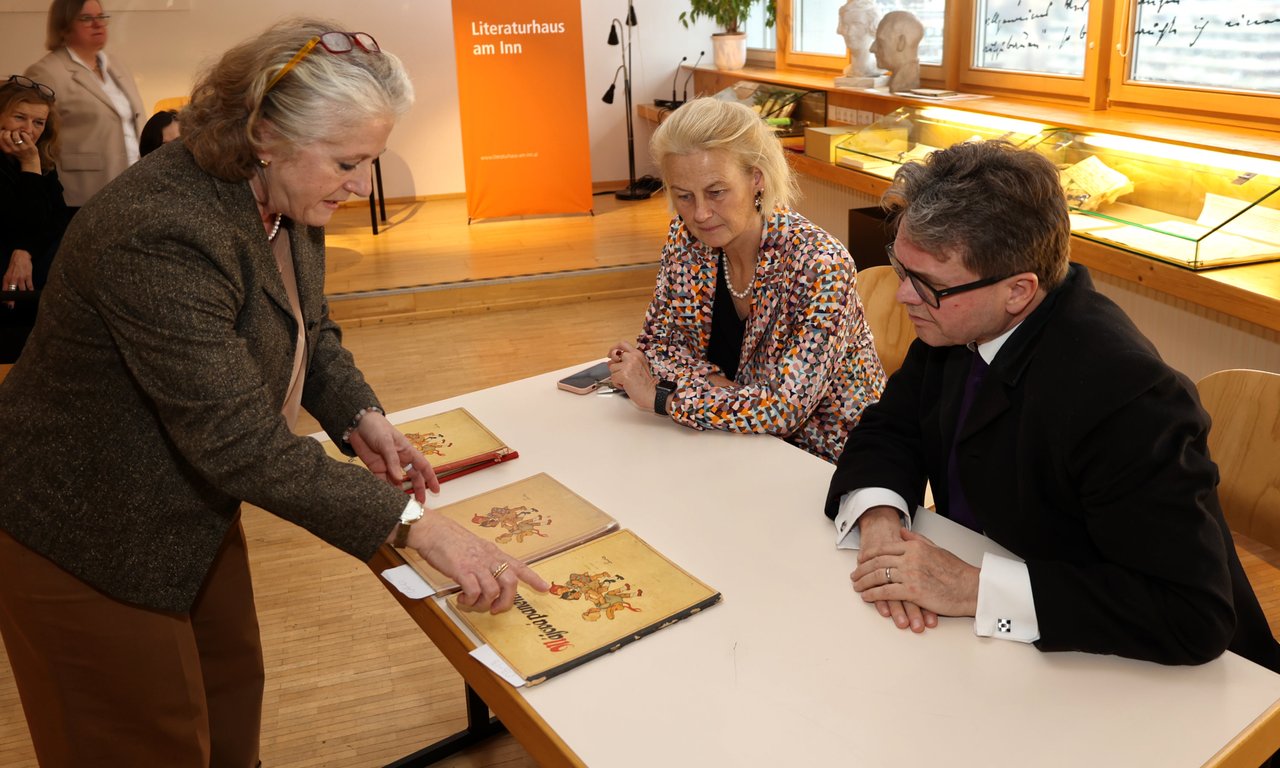 Zwei Personen sitzen an einem Tisch und betrachten Bücher, die von einer dritten, am Tisch stehenden Person präsentiert werden.
