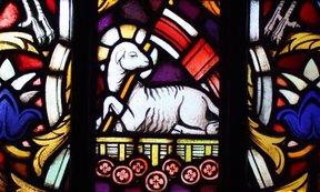 Zu sehen ist ein buntes Kirchenfenster. Darauf abgebildet ist ein weißes Lamm mit Kreuzesfahne.