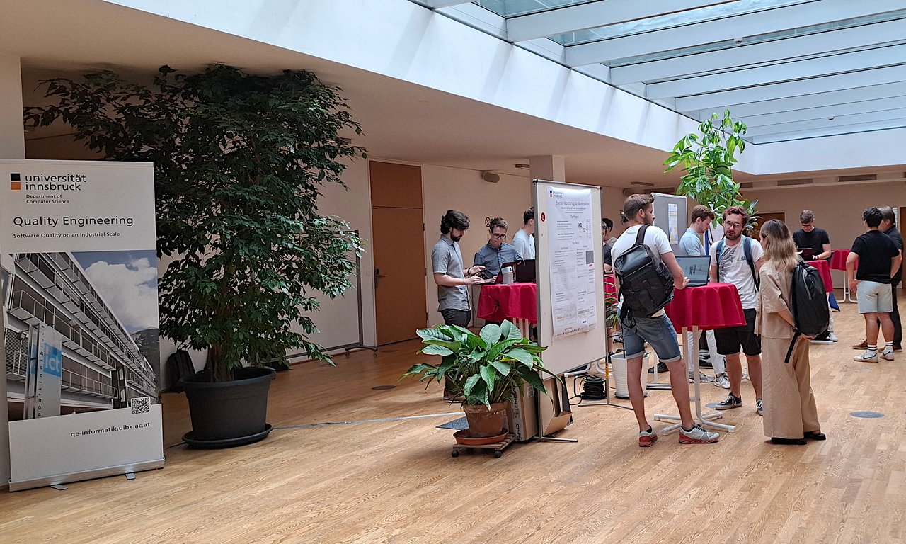 das das Foyer vom ICT-Gebäude, links ein roll up von der Fachgruppe Quality Engineering und in der Mitte haben alle Teams ihre Poster von ihrem Projekt ausgestellt und stehen an Tischen und unterhalten sich