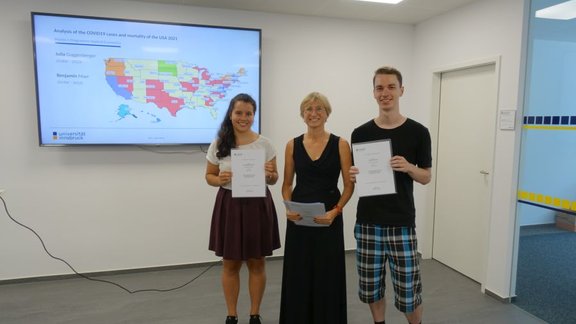 Two students holding a certificate, with Joanna in the middle, stand in front of a screen showing a map of the US. It is a slide on their project "Analysis of the Covid-19 cases and mortality of the USA 2021".