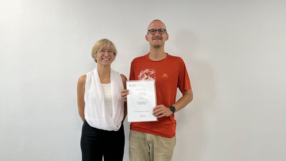 Joanna and a student standing next to each other in front of a white wall. The student holds a certificate in his hands.