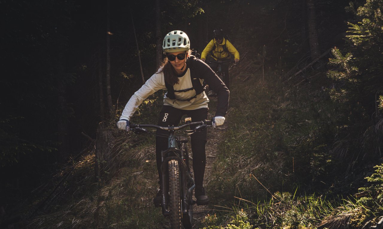 Frau fährt mit Mountainbike durch den Wald. Im Hintergrund eine zweite Person auf dem Fahrrad.