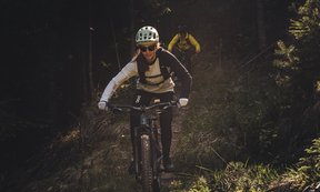 Frau fährt mit Mountainbike durch den Wald. Im Hintergrund eine zweite Person auf dem Fahrrad.