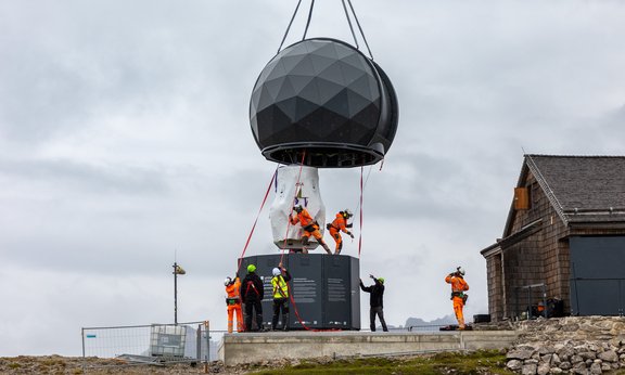 Eine schwarze Kuppel hängt an Gurten und schwebt dicht über dem Unterbau. Montagearbeiter ziehen die Kuppel in Position.