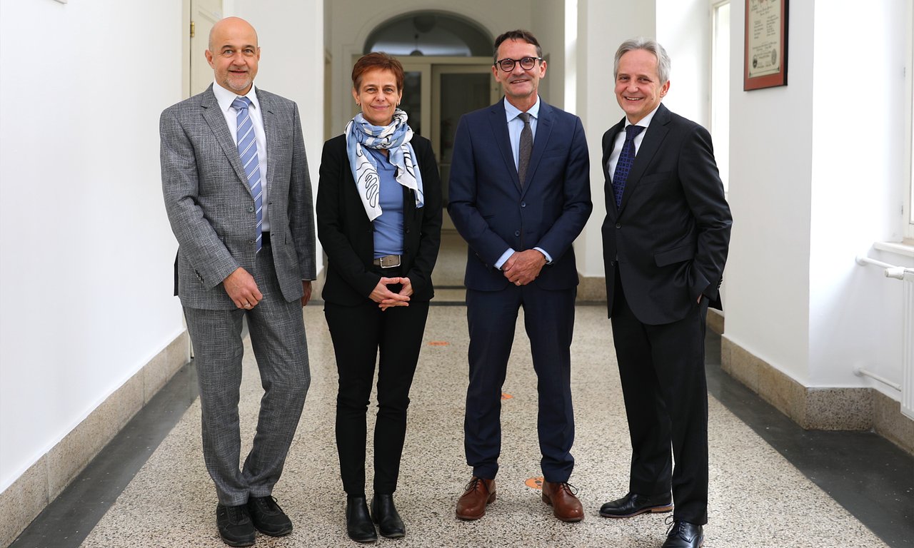 Vier Personen stehen in einer Reihe in einem Gang und blicken in die Kamera