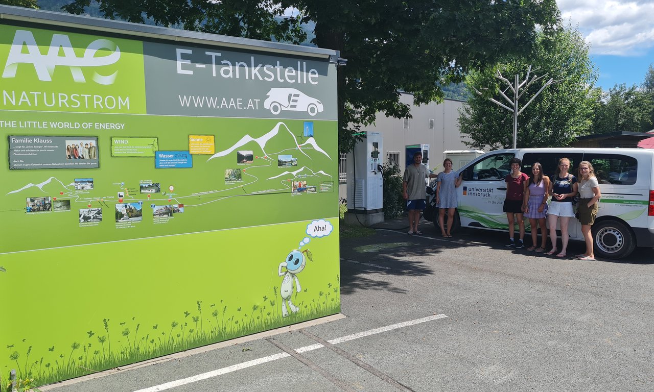 Im Vordergrund ein großes Schild einer E-Tankstelle, dahinter Studierende und ein Kleinbus mit Uiversitätsbeklebung.