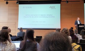 Zu sehen sind die Teilnehmer und Teilnehmerinnen der Tagung „Lehren und Lernen im digitalen Zeitalter“. Sie befinden sich in einem Vortragssaal. Sie folgen einer Präsentation.