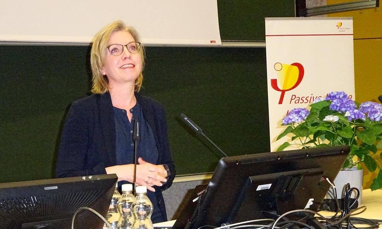 Eine Frau steht vor Bildschirmen, hinter ihr eine Tafel, neben ihr Blumen.