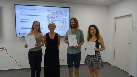 Four people standing in front of a screen with a slide on the project "Wikipedia Gender Content Gap". Joanna, in a black dress, is surrounded by three students to her left and right, holding certificates.