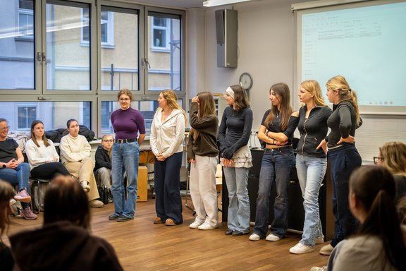 7 Schülerinnen stehen inmitten eines Klassenzimmers, eine von ihnen spricht. Rund herum sitzen Zuschauerinnen.