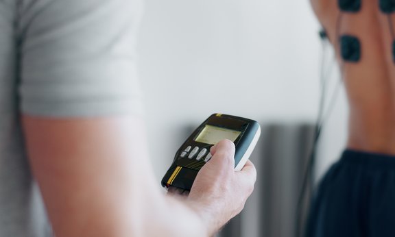 Person hält Analysegerät, im Hintergrund ist eine Person zu sehen, die an Sensoren angeschlossen ist