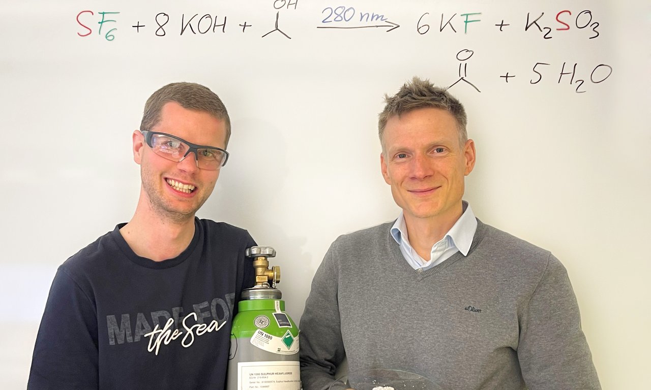 Zwei Personen mit einer Gasflasche und einem Glasteller mit weißem Pulver in der Hand, dahinter ein Whiteboard mit chemischen Formeln.