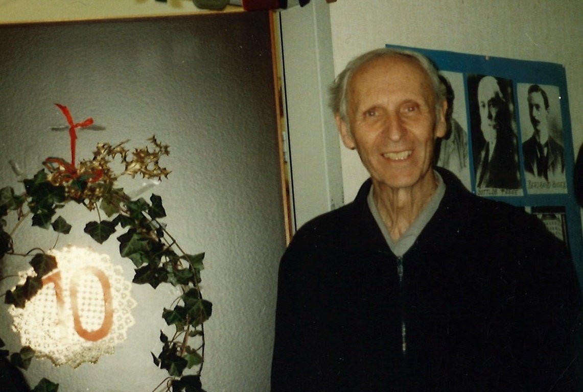 Ein Mann steht vor einer offenen Tür, seitlich hinter ihm ist ein Gang mit Fotos an der Wand zu sehen.