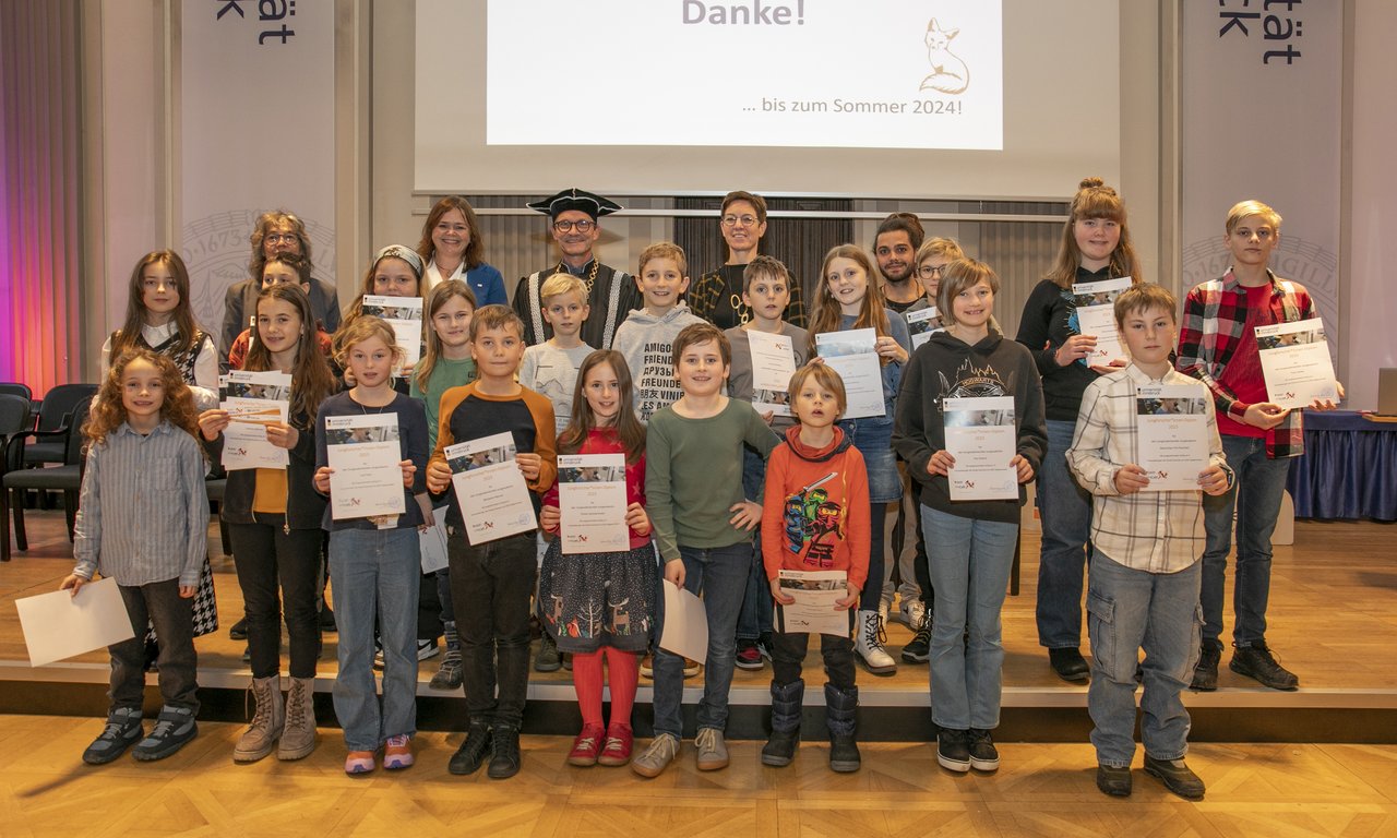 Gruppenfoto auf der Bühne der Aula, die Kinder mit ihren Diplomen und Vertreter:innen der Hochschulen sind zu sehen.