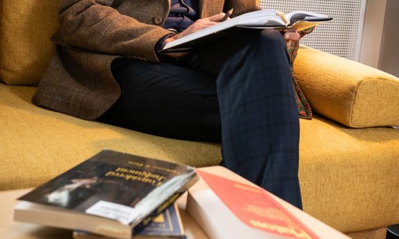 Mann sitzt mit verschränkten Beinen auf Sofa, ein Buch auf den Knien