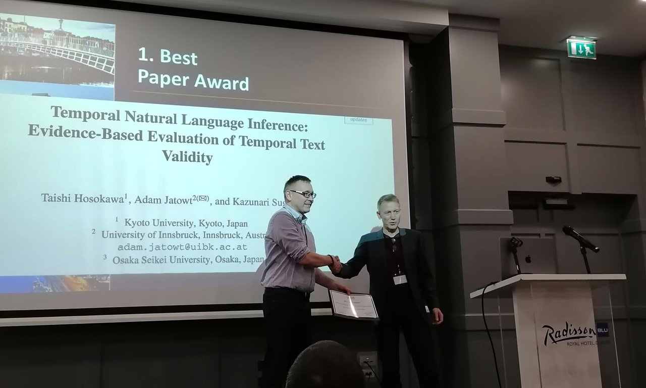 Zwe Männer schütteln sich auf einer Bühne die Hand, dahinter sind Titel und Abstract eines wissenschaftlichen Papers projiziert.
