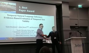 Two men shake hands on a stage, behind which the title and abstract of a scientific paper are projected.