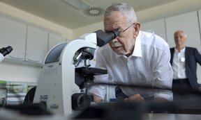Alexander Van der Bellen schaut durch ein Mikroskop, im Hintergrund ist ein weiterer Mann zu sehen.