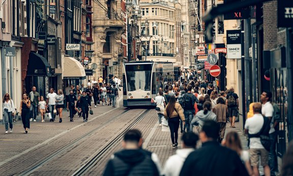 Symbolbild: Eine Straßenszene aus Amsterdam, zu sehen sind ein Straßenzug, viele Menschen auf den Seiten und eine Straßenbahn auf der Straße.