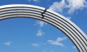 Ein halber Bogen aus weißem Stein vor einem blauen Himmel mit Wolken, auf dem Bogen ist ein aufgeklebter Riss zu sehen.