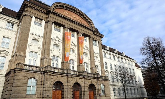 Zwei orangefarbene Fahnen hängen an der Fassade des Hauptgebäudes der Uni Innsbruck.