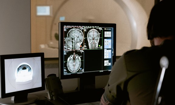 Arzt schaut auf einen Bildschirm mit Gehirn-MRT, im Hintergrund ein MRT-Gerät.