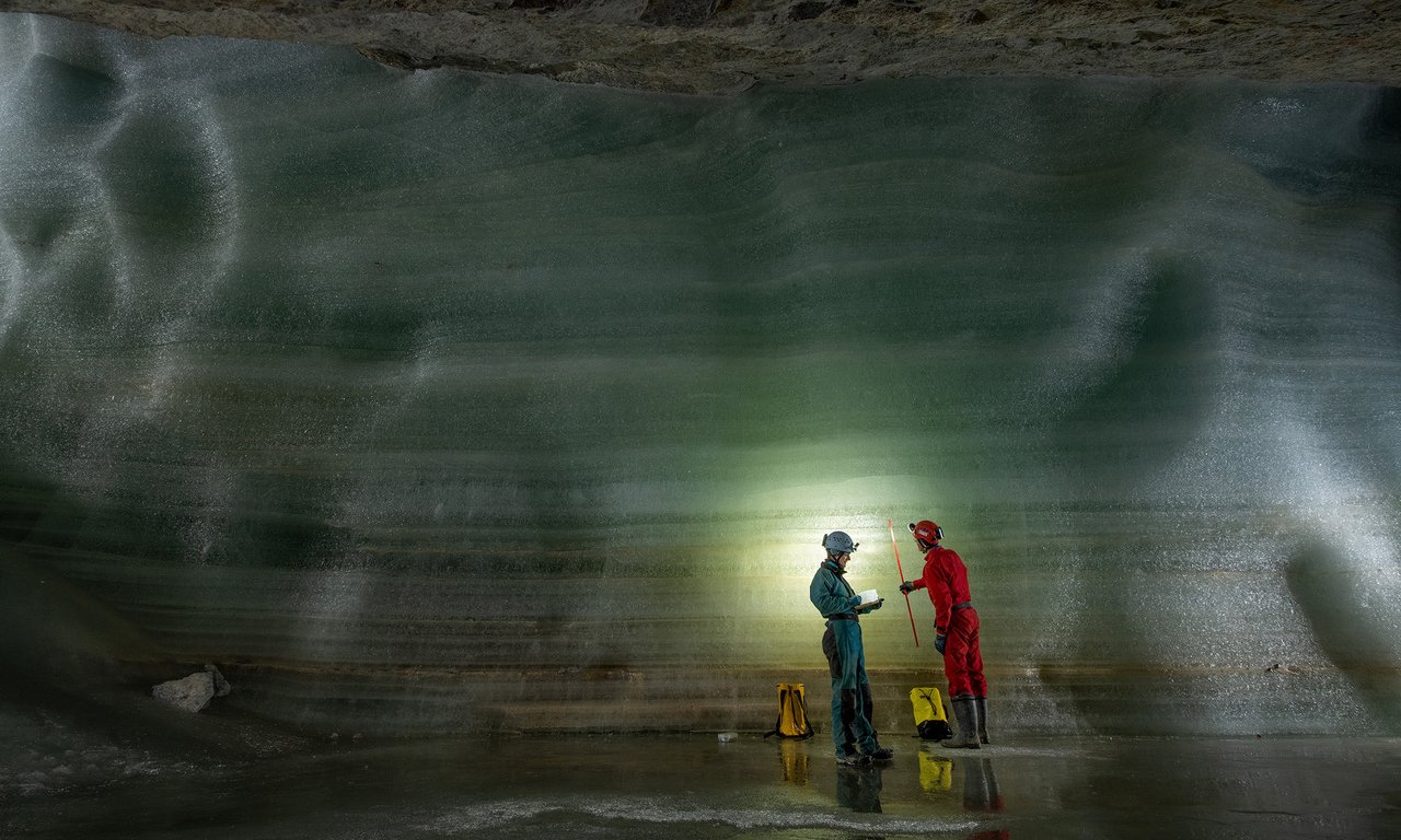 Zwei Personen in Overalls und mit Helmen stehen vor einer Eiswand, es ist dunkel, ein Licht-Spot beleuchtet eine Stelle.