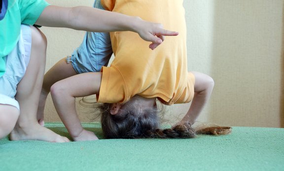 Ein Kind versucht einen Kopfstand zu machen, eine Hand deutet mit dem Zeigefinger nach rechts