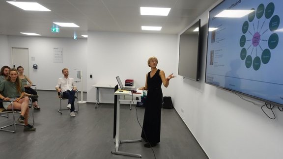 Joanna is standing at a podium, looking and showing towards a screen behind her. You can also see four people in the audience, listening to her.