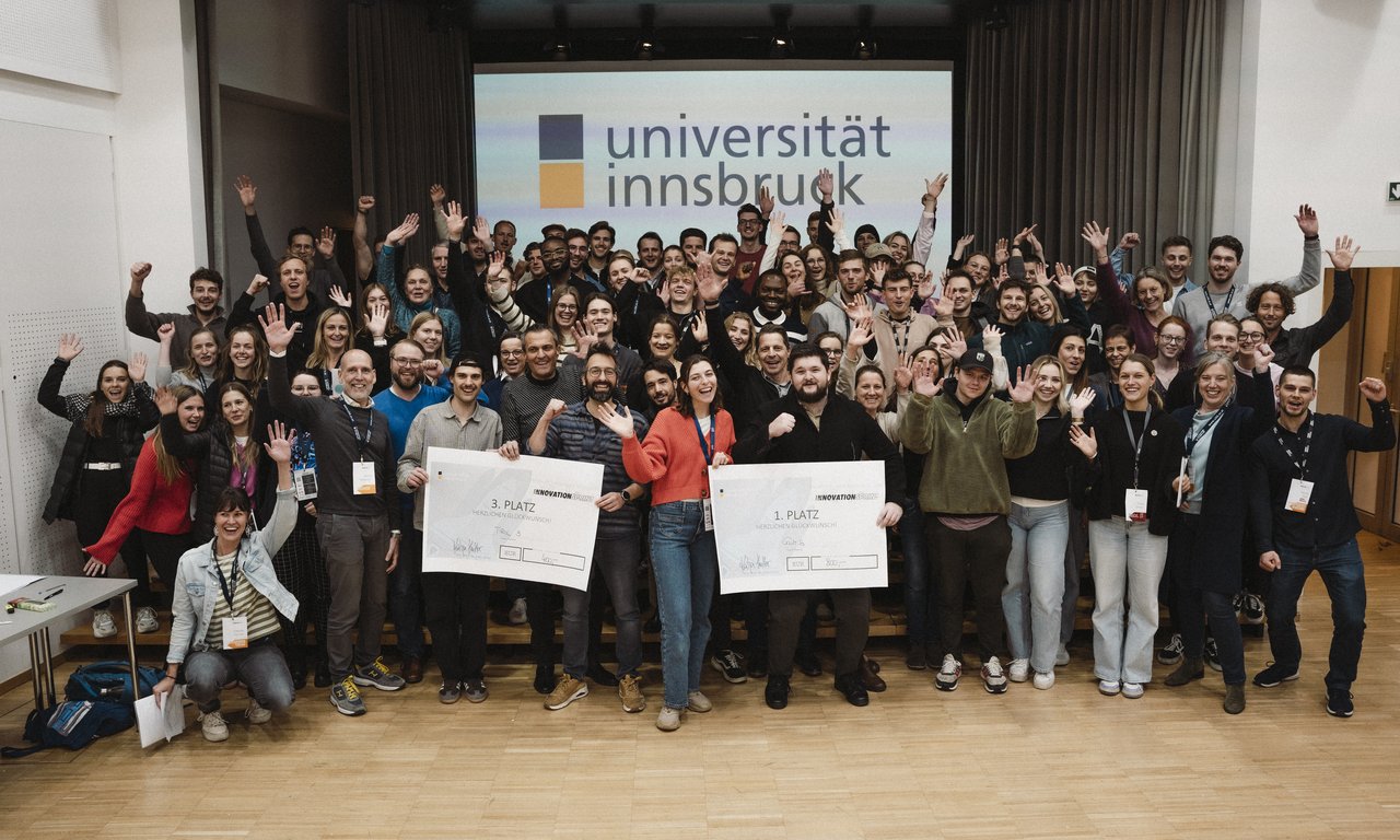 Dutzende von Menschen jubeln in die Kamera, im Hintergrund das Logo der Universität Innsbruck
