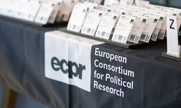 A registration desk belonging to the European Consortium for Political Research (ECPR), covered with a black cloth, on which numerous name badges are arranged in alphabetical order.