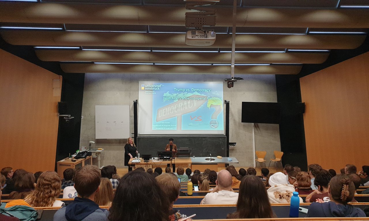 Ein gut gefüllter Hörsaal von einer Position ganz hinten im Raum. Im vorderen Bereich stehen Cornelia Klecker und Lore Hayek.