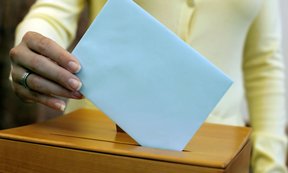 Eine Frauenhand wirft ein Kuvert in eine Wahlurne