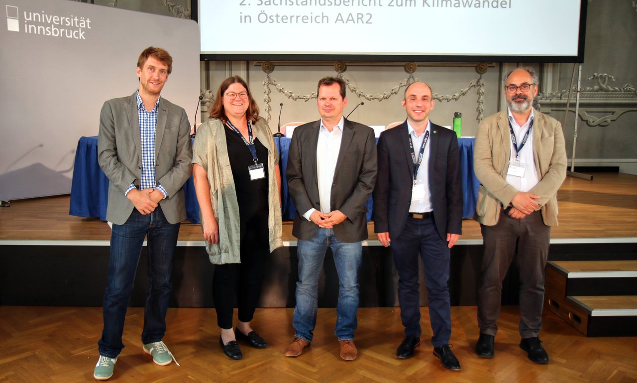Fünf Personen stehen vor einer Bühne, im Hintergrund ist ein Tisch für Pressegespräch zu sehen und „2. Sachstandsbericht zum Klimawandel in Österreich AAR2“ ist an eine Wand dahinter projiziert