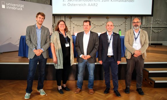 Fünf Personen stehen vor einer Bühne, im Hintergrund ist ein Tisch für Pressegespräch zu sehen und „2. Sachstandsbericht zum Klimawandel in Österreich AAR2“ ist an eine Wand dahinter projiziert