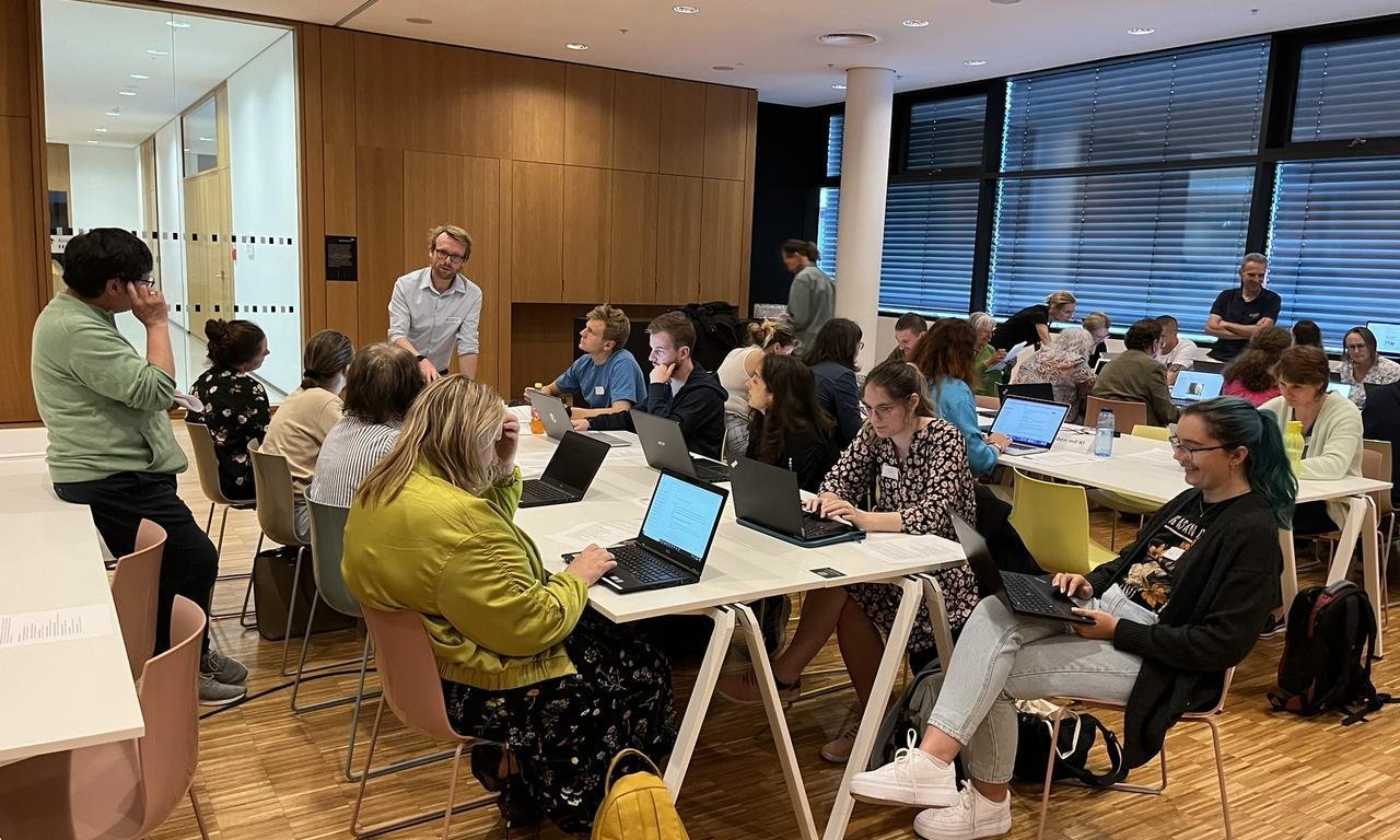 Blick in einen Vortragsraum: Zahlreiche Menschen arbeiten an drei langen Tischen auf ihren Laptops und diskutieren, drei Männer stehen jeweils an einem der Tische und beraten.