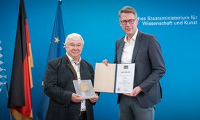Rainer Blatt und Markus Blume halten die Urkunde und die Bronzetafel in der Hans und blicken in die Kamera, im Hintergrund die deutsche und EU-Fahne.