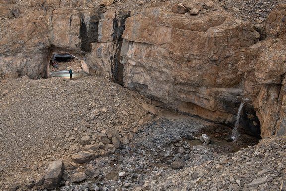 Blick auf steiniges Gelände mit einem kleinen Wasserfall und Höhleneingang im Hintergrund.