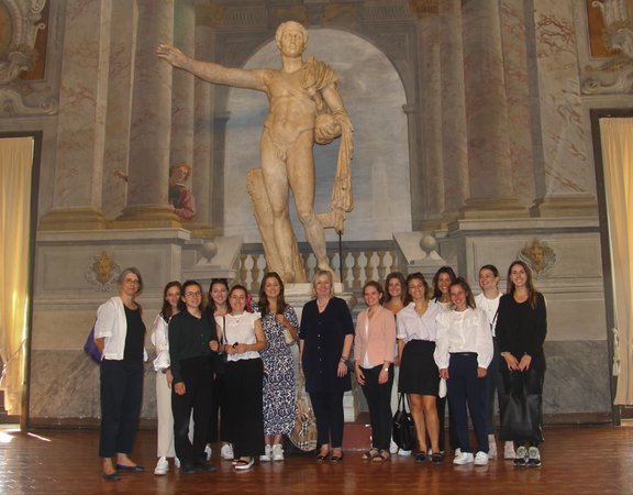 14 Frauen stehen für ein Gruppenfoto vor einer antiken Statue im Innenbereich eines Gebäudes zusammen.