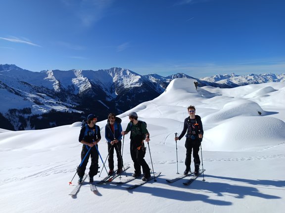 Auf dem Bild sind vier Mitglieder des Instituts zu sehen. Sie stehen auf der Skipiste. Die Sonne scheint und die Personen sind sichtlich guter Laune.