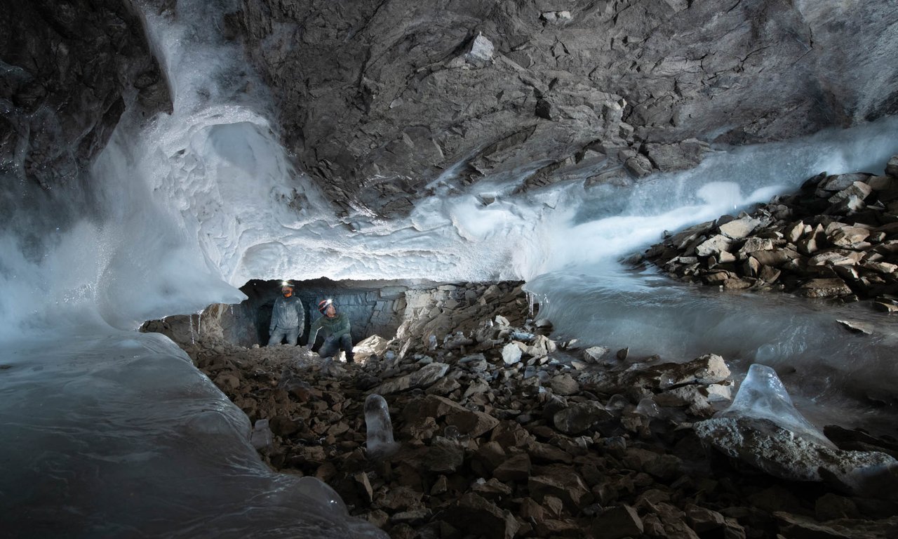View of the entrance to a cave with dark rock formations, partial ice formations, and two scientists.