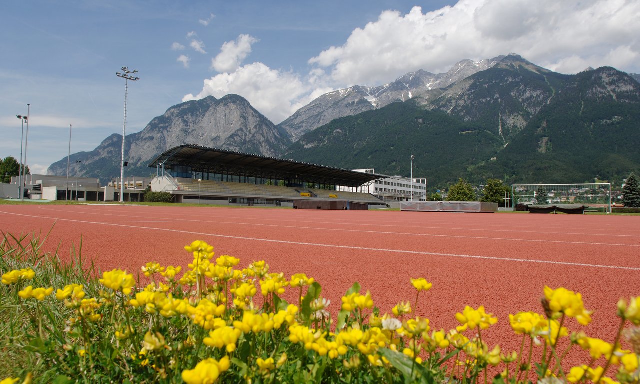 Sportcampus der Universität, zu sehen sind im Vordergrund Blumen vor einer Leichtathletikbahn im Freien, dahinter Gebäude.