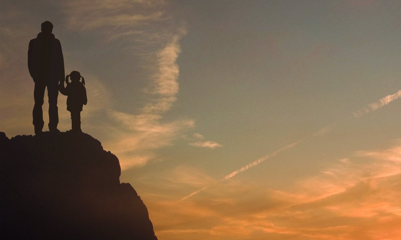 Eine große und eine kleine Person stehen auf einem Felsen und blicken bei Abendstimmung Richtung Horizont