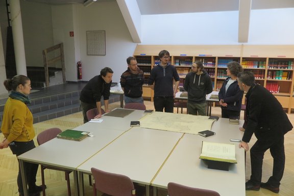 Seven people arranged around a big table discussing a map and books.
