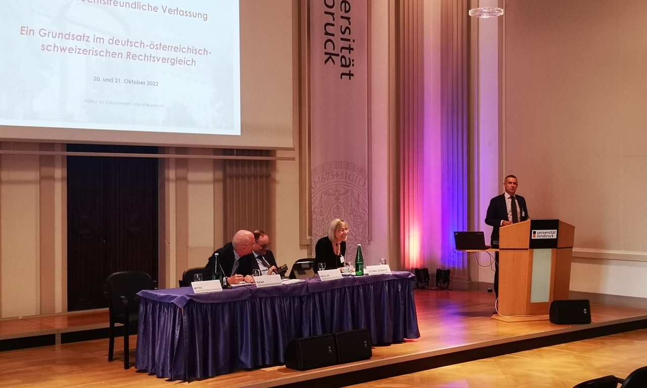 Drei Personen auf einem Podium, daneben spricht jemand an einem Rednerpult.
