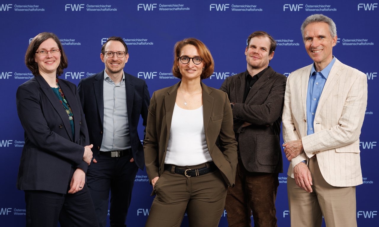 Fünf Personen stehen in einer Reihe und blicken in die Kamera, im Hintergrund eine blaue Leinwand.