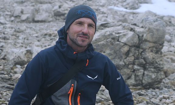 Das Bild zeigt Thomas Klotz im Portrait. Er trägt Outdoor-Kleidung und ist im Gelände. Im Hintergrund sind Felsen zu sehen.