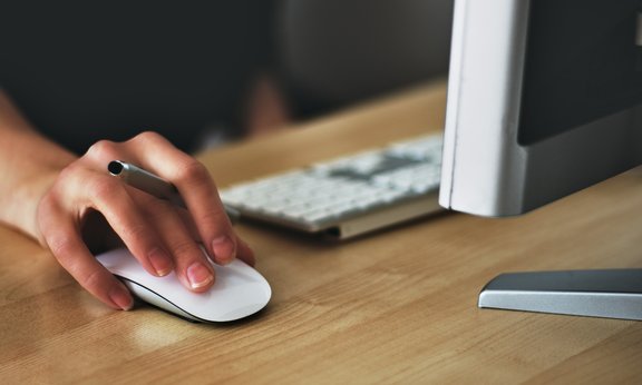 Ein Mann hält eine Computermaus in der Hand, die Tastatur und ein Bildschirm von hinten sind auch zu sehen.